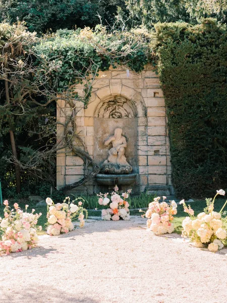 Ceremony backdrop with hydrangea and rose ground florals framing a stone wall fountain and statue along a gravel garden aisle