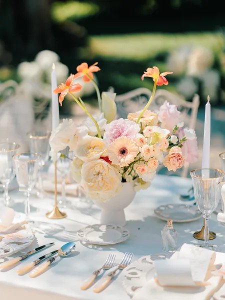 Reception tablescape with a wedding table centerpiece of peach and blush flowers in a white vase, surrounded by taper candles on a garden table with white chairs