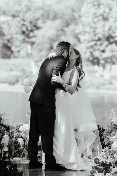 Wedding kiss portrait of bride and groom kiss in a garden ceremony, her veil and sleeveless dress visible beside floral arrangements
