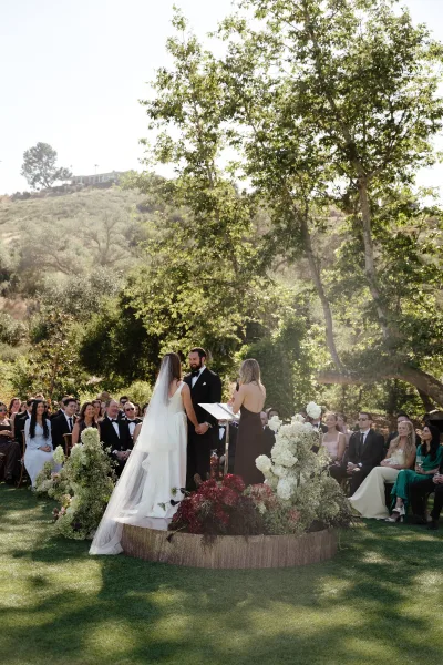 Ceremony moment at an outdoor wedding ceremony as bride and groom hold hands during vows, with veil and acrylic podium on a lawn with guests