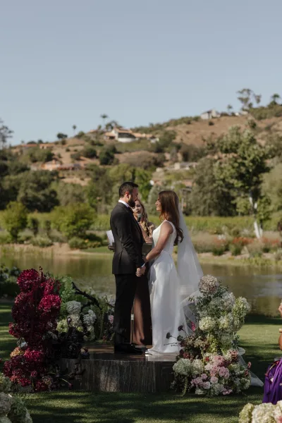 Wedding vows as bride and groom hold hands under a floral arch with hydrangeas and roses beside a lake, officiant on microphone