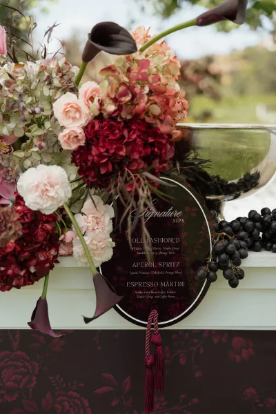 Wedding bar menu and signature drinks sign on a round acrylic board with burgundy florals, grapes, and glass punch bowl outdoors