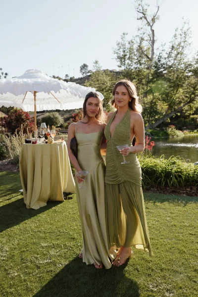 Bridesmaid portrait in sage green bridesmaid dresses holding martini glasses by a high-top table under a white patio umbrella by the pond