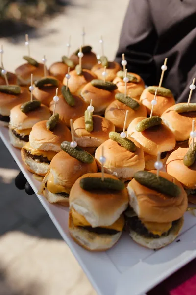 Wedding sliders on a serving tray with mini burgers, pickle garnish, and appetizer picks, carried by a server on outdoor pavement