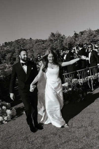 Wedding recessional with bride and groom walking the aisle, her long veil flowing as guests cheer beside florals on an outdoor lawn