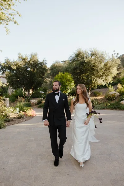 Couple portrait of bride and groom walking hand in hand on a stone garden path, she holds a calla lily bouquet in a sleek dress