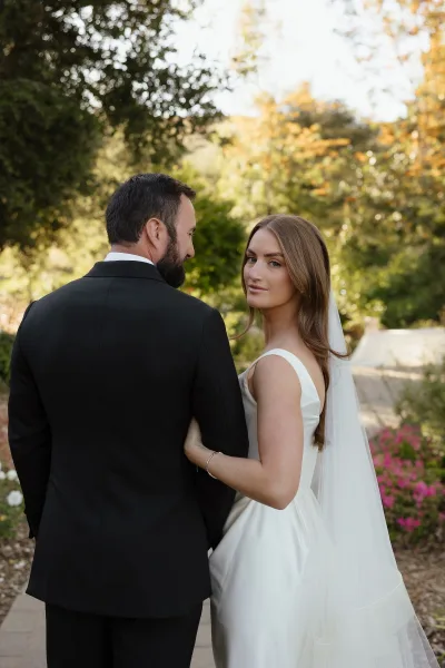 Couple portrait of newlyweds walking away, bride looking back in veil and satin dress holding groom’s arm on a sunlit garden path
