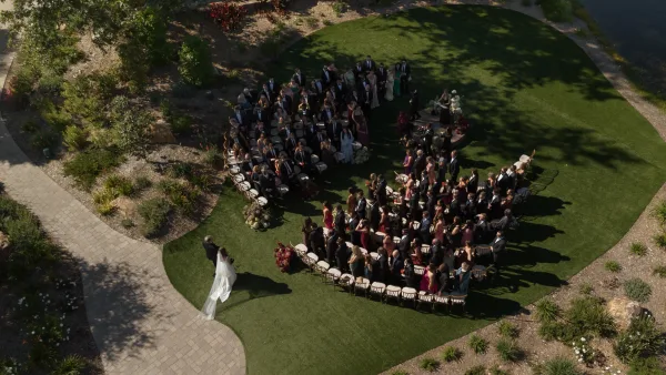 Outdoor wedding ceremony with curved chair rows as a bride with long veil walks the grass aisle toward a floral arch on a garden lawn