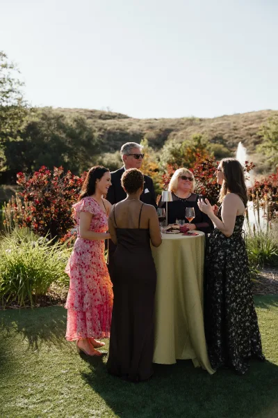 Cocktail hour guests mingle around a standing table with wine glasses and sliders, chatting on a lawn with fountain and water views.