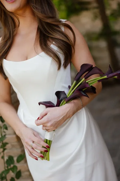 Bridal portrait of a bride holding bouquet with calla lily bridal bouquet, showing rings and bracelet against stone wall and greenery backdrop