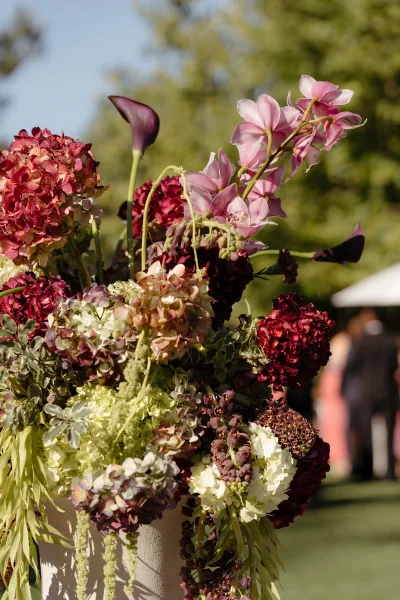 Wedding floral arrangement of hydrangea and orchids on a pedestal with greenery, set on an outdoor lawn with trees and blurred guests