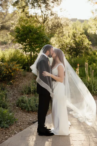 Wedding kiss as bride and groom kiss under a flowing veil on a sunlit garden stone walkway, groom in black tuxedo