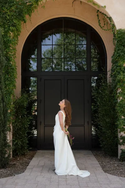 Bridal portrait of a bride in a simple wedding dress holding a purple calla lily bouquet by black doors under an ivy-covered arch
