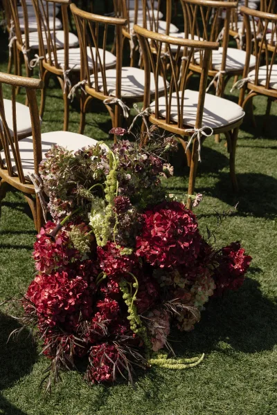 Ceremony aisle decor with hydrangea aisle flowers in a grounded floral cluster beside wood chairs with white cushions and ribbon ties on a sunny lawn