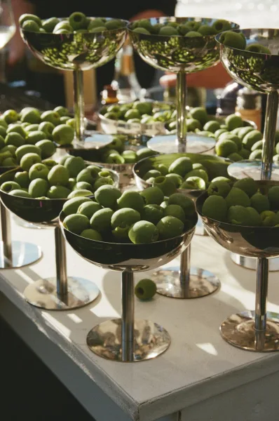 Cocktail hour snacks at a wedding olive bar, green olives in metal coupe glasses and bowls on a white bar top with blurred guests