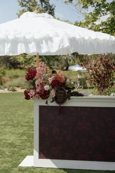 Cocktail bar setup with a mobile bar cart, cocktail menu sign, and white patio umbrella on a lawn with garden trees behind