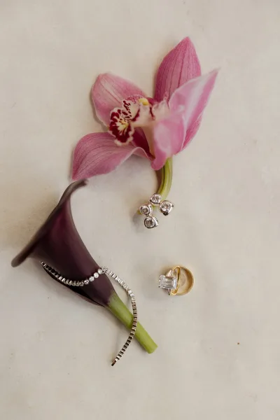Wedding rings with emerald cut engagement ring and gold band on white fabric, accented by diamond bracelet, earrings, and orchid blooms