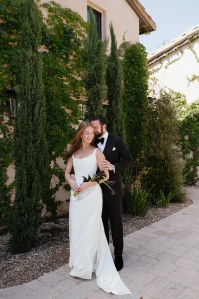 Couple portrait of bride and groom embrace, she holds black calla lily bouquet with ribbon beside an ivy-covered stucco wall on stone walkway
