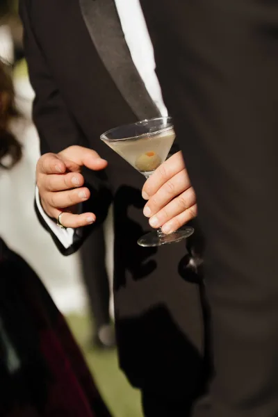 Wedding cocktail martini wedding drink held by a groom in a tuxedo, olive garnish in the glass and ring on hand, greenery behind