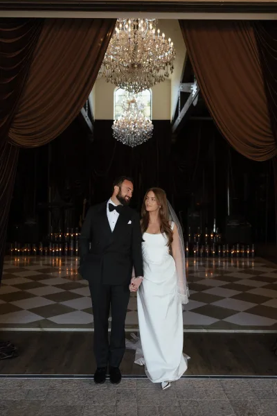 Couple portrait of bride and groom holding hands, her long veil flowing beside his tuxedo under chandelier in a moody ballroom