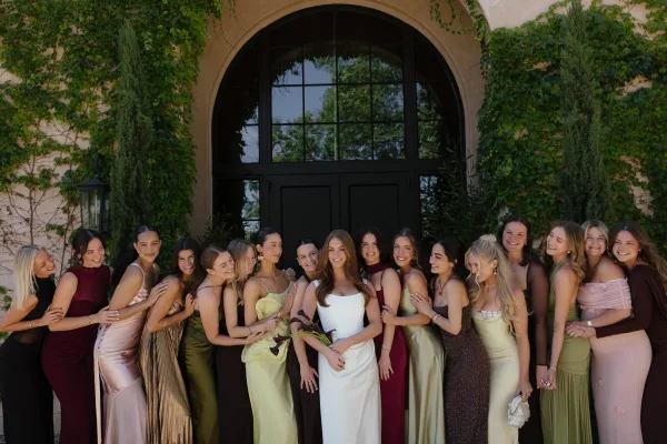 Bridesmaid group photo of the bride with bridesmaids in satin dresses, holding a calla lily bouquet by an ivy-covered arched doorway