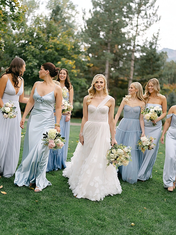 Bride with bridesmaids walking together, all holding bouquets, with blue satin bridesmaid dresses on a green lawn with mountains behind