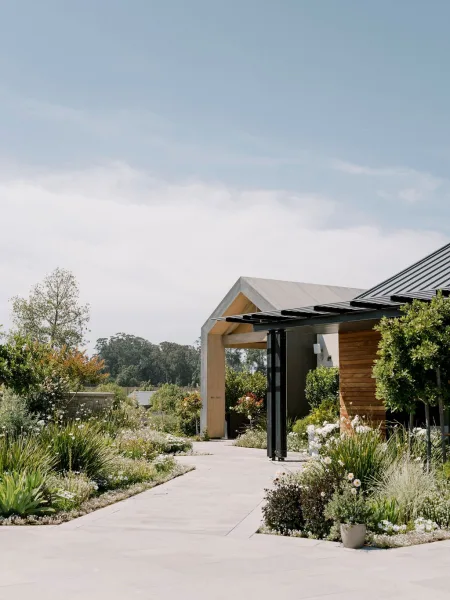 Wedding venue exterior with modern barn architecture, glass doors and wood siding along a concrete walkway with potted flowers under blue sky
