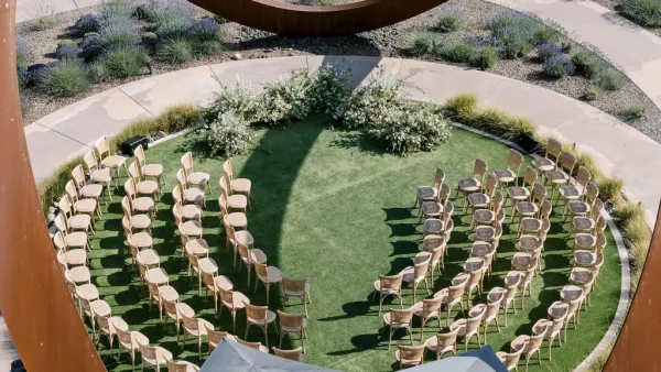 Ceremony setup with outdoor ceremony seating in a semicircle of wood chairs around a grass aisle, with white-green florals and curved metal structure on a circular lawn