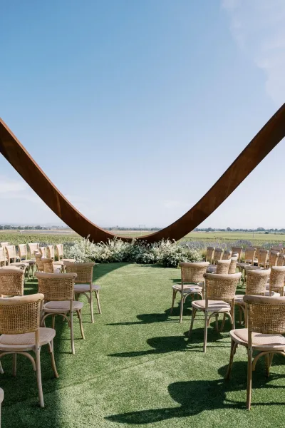 Ceremony setup with outdoor wedding ceremony setup featuring a curved wood arch, white florals and greenery, wicker chairs, and green aisle runner in an open field