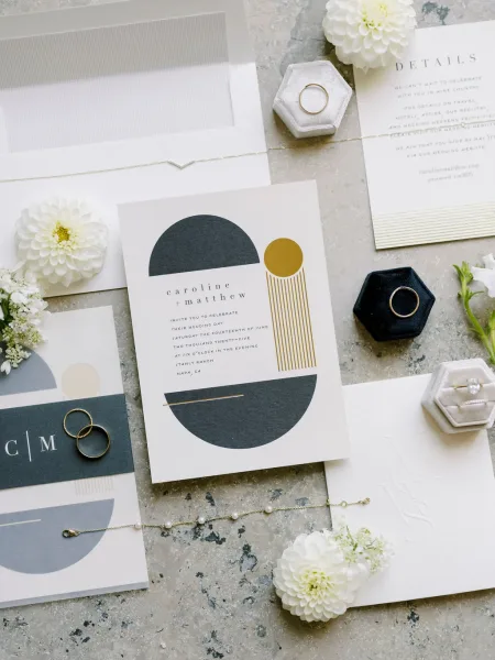 Invitation suite of modern wedding invitations with envelopes, wax seal, rings, pearl bracelet and white flowers arranged on stone surface