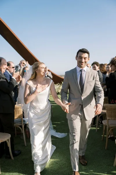 Wedding recessional as bride and groom walk hand in hand down the outdoor aisle, bride laughing in veil while guests cheer under blue sky by a metal sculpture