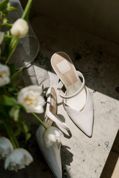 Bridal shoes in a sunlit flat lay, white wedding heels with slingback straps beside flowers and a glass vase on stone surface