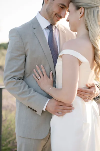 Couple portrait of bride and groom close up, embracing with foreheads touching in a sunset-lit field, ring and bracelet visible