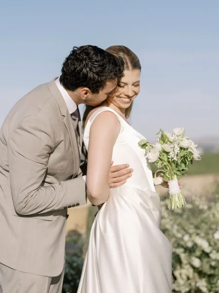 Couple portrait of groom kissing bride’s cheek as they embrace, bride holding a white rose bouquet in a field under blue sky