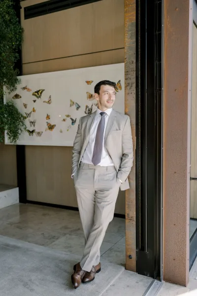 Groom portrait in a light gray suit with hands in pockets, looking away by an indoor venue entrance with butterfly wall art