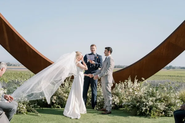 Wedding vows as bride and groom hold hands, officiant reads at a sculptural altar in a windy open field under blue sky