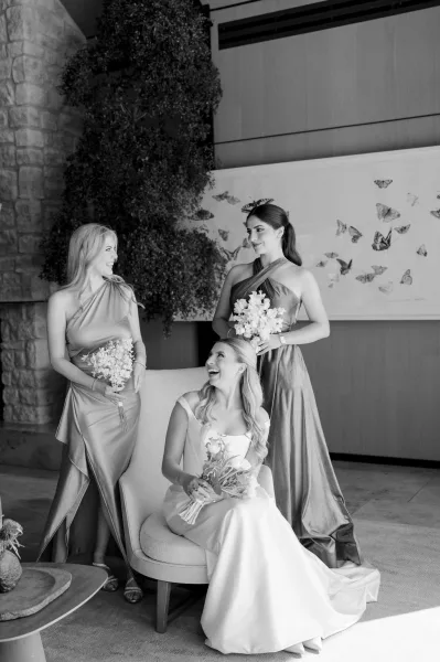 Bridesmaid portrait with bride and bridesmaids in satin dresses holding white bouquets, gathered by a stone wall and indoor tree in a lounge setting