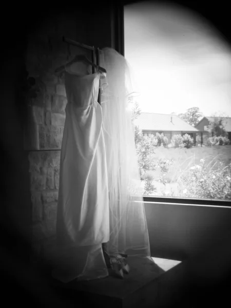 Wedding dress with veil hanging by a window on a dress hanger, soft light on a stone wall with barn and trees beyond