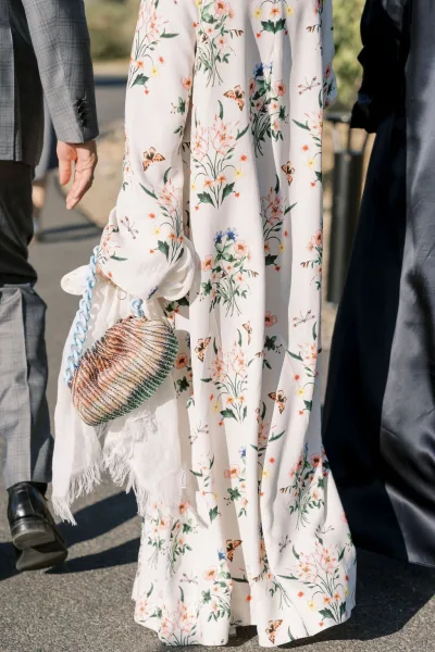 Wedding guest outfit featuring a floral maxi dress with beaded clutch and fringe shawl beside men in suits on outdoor pavement by chairs