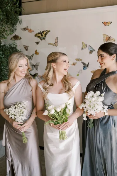 Bridesmaid portrait of a bride with bridesmaids holding white tulip bouquets, wearing mismatched dresses, posed by a butterfly wall backdrop indoors