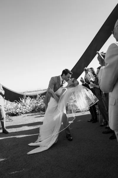 Wedding kiss as bride dips back in strapless dress with long veil blowing, groom in light suit on outdoor terrace by guests clapping
