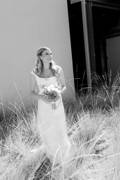 Bridal portrait of a smiling bride holding bouquet in an off-the-shoulder wedding dress, standing by tall grass near a doorway wall