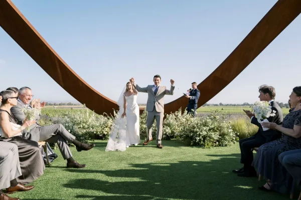 Recessional moment as bride and groom cheering while walking up the aisle, veil and white bouquet in an open field under a wooden arch
