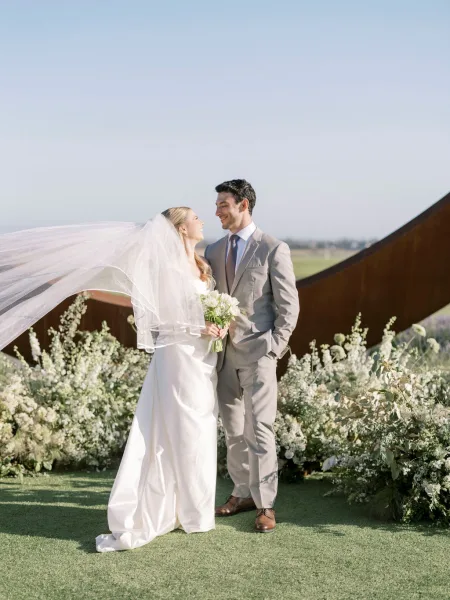 Couple portrait of bride and groom smiling as her veil blows, holding a white bouquet on a lawn with blue sky and sculptural metal wall