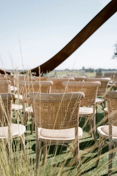 Ceremony seating with outdoor wedding chairs in natural wood and woven cane, set on grassy aisle in open meadow with distant hills and sky