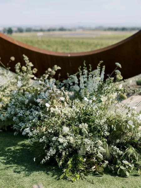 Wedding ceremony altar with white and green florals and greenery around a curved backdrop panel on a grassy field with distant hills