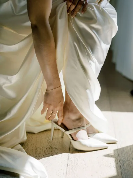 Bridal shoes, white wedding heels with pointed toes and ankle straps, as bride adjusts them on a sunlit wood floor with ring and bracelet