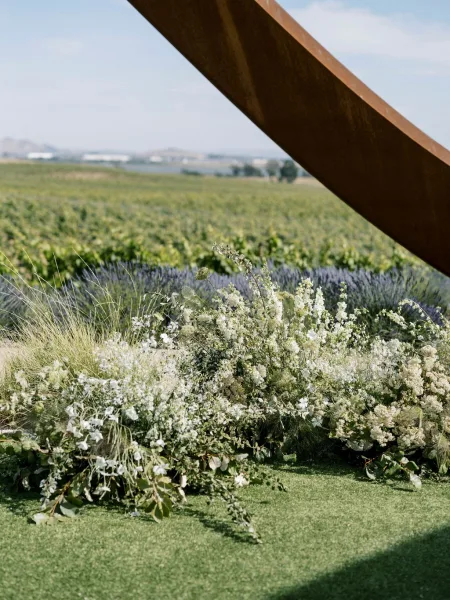 Wedding altar flowers on a modern metal arch with white blooms, wildflowers and greenery on a grass lawn by a lavender field and hills