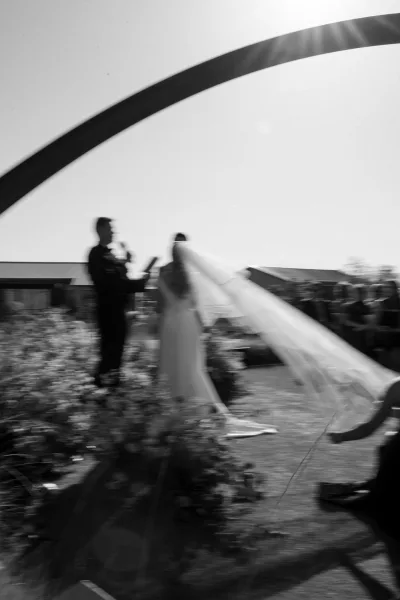Ceremony moment at an outdoor wedding ceremony as bride and groom stand at the altar under a circle arch, veil blowing as guests watch