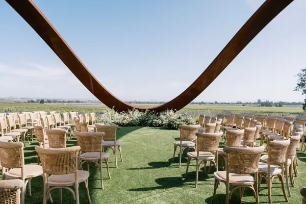 Ceremony setup for an outdoor wedding ceremony with modern wood arch, wicker chairs, green aisle runner, and white florals in an open field